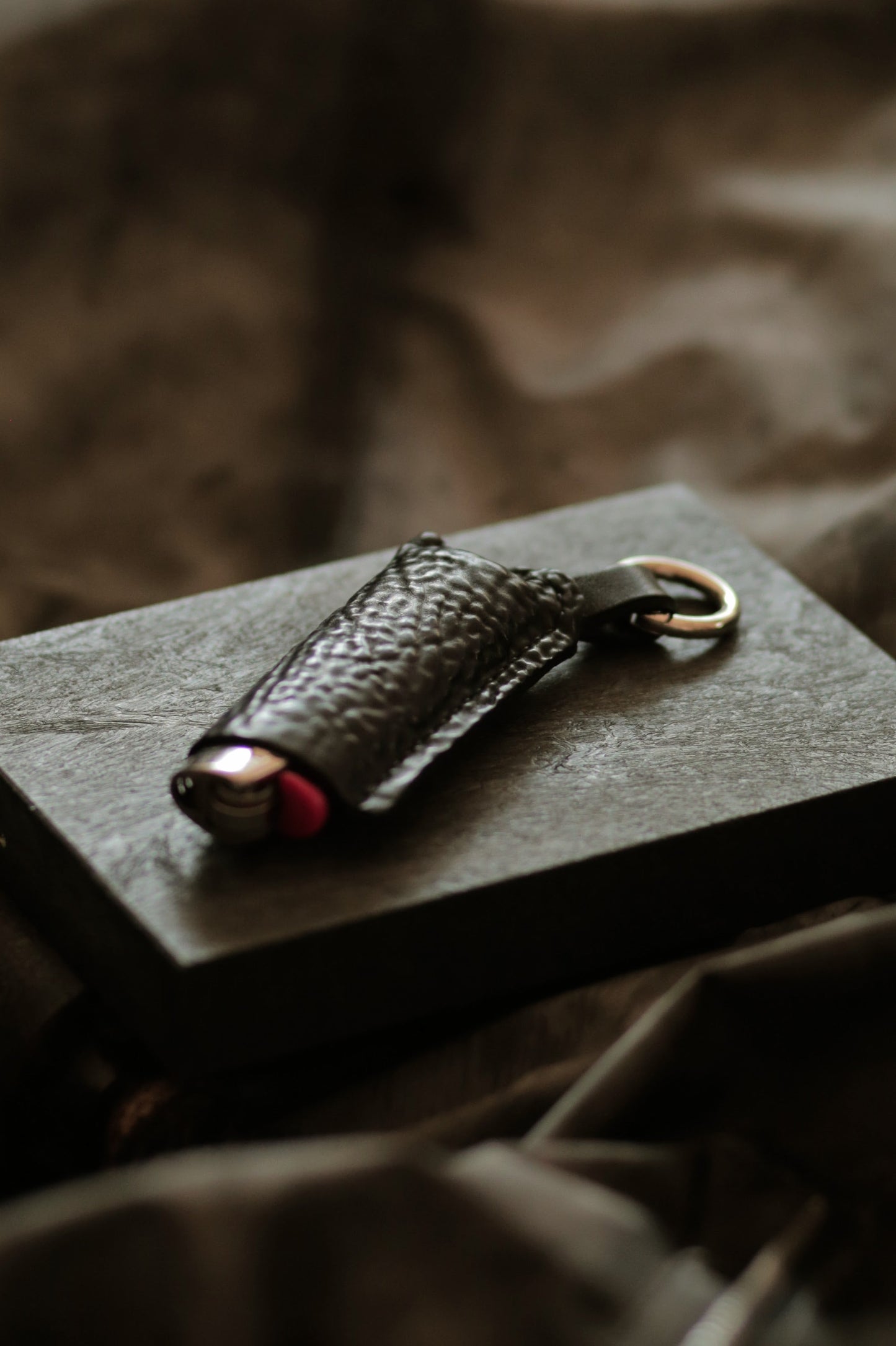 Lighter with a horse culatta leather case resting on a dark textured surface with metal keyring attached.