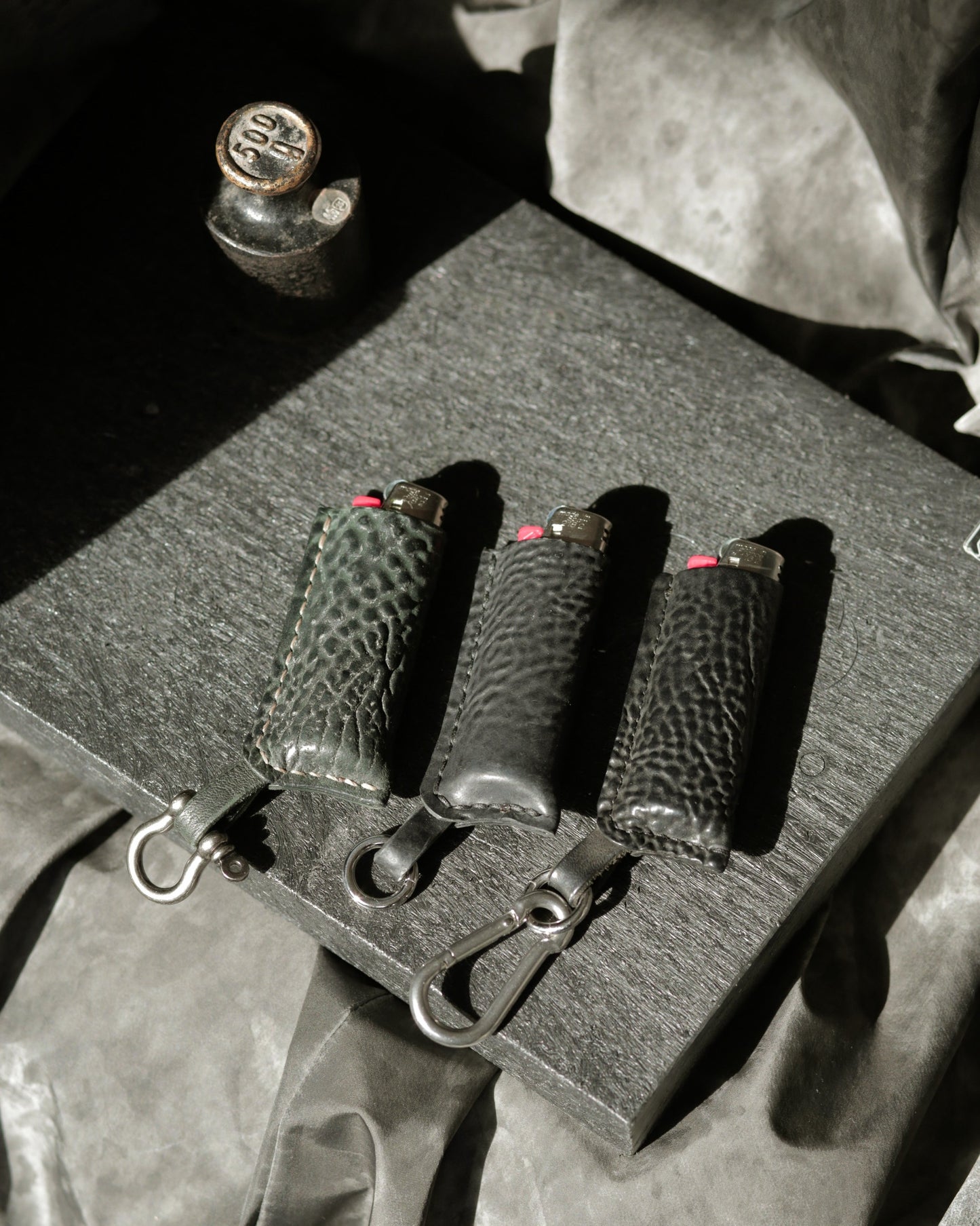 Lighter with a case in dark green and two black horse culatta leather products displayed on black background.